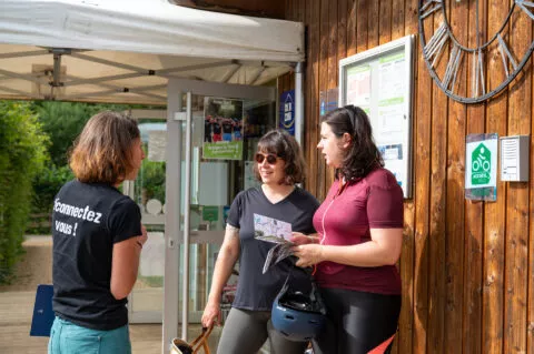 Deux femmes cyclotouristes sont accueillies par une autre femme à l'entrée d'un camping labélisé Accueil Vélo